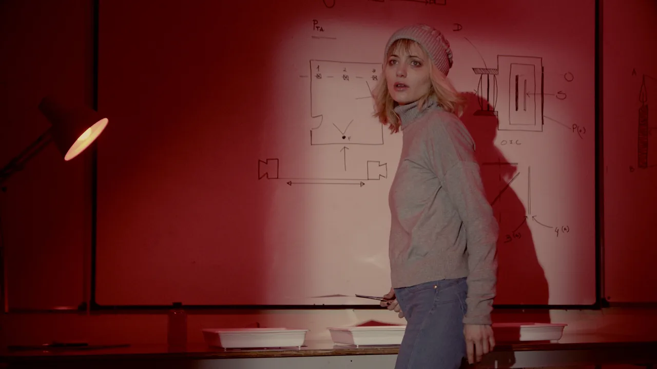 A woman in a knitted hat and gray sweater stands before a whiteboard under red light